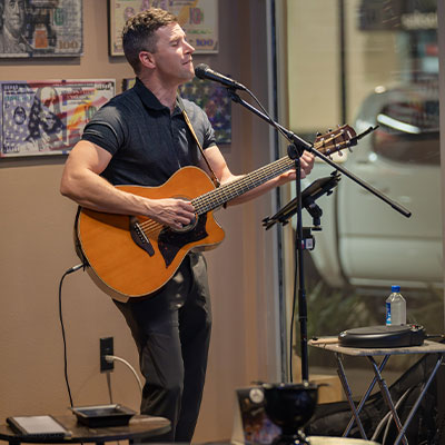 A person stands and sings into a microphone while playing an acoustic guitar in a room decorated with wall art. A music stand, a bottle of water, and audio equipment are visible.