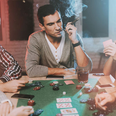 A group of people sit around a table playing poker. One man in the center is smoking a cigar and holding playing cards. Poker chips, cards, and drinks are on the table.